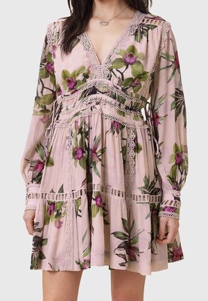 Vrouw die een lichtroze bloemenjurk draagt met paarse en groene bloemen, kanten randen, lange mouwen en een V-hals.