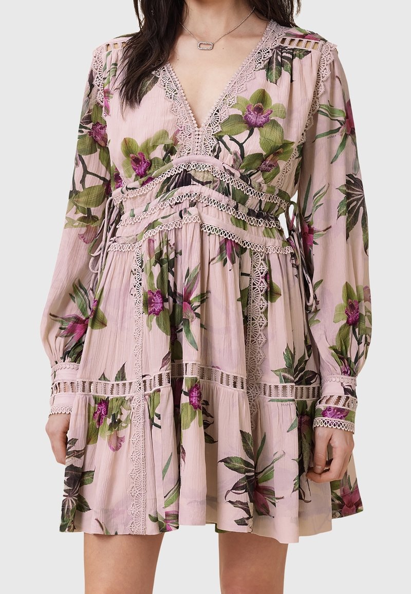 Femme portant une robe fleurie rose clair avec des fleurs violettes et vertes, détails en dentelle, manches longues et col en V.