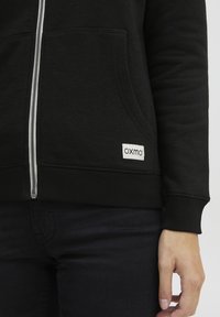 Crna zip-up dukserica od mekanog materijala, s prednjim džepovima, rebrastim manžetama i bijelom etiketom s natpisom "OXMO" na donjem lijevom dijelu.