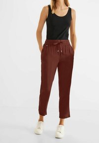 Kvinna som bär en svart ärmlös topp, rödbruna dra-på-byxor och vita sneakers, stående med händerna i fickorna på en vit bakgrund.