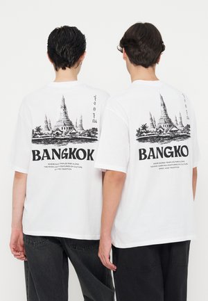 Δύο άτομα φοράνε λευκά T-shirts με μαύρο σκίτσο ενός ναού στην Μπανγκόκ και κείμενο για τον πολιτισμό και την παράδοση στο πίσω μέρος.