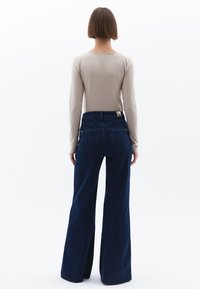 Béžový top s dlouhým rukávem, který je kombinován s vysokými tmavě modrými kalhotami s širokými nohavicemi, jež mají dvě zadní kapsy a jemnou texturu látky.