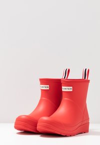 Botas de lluvia de goma rojas con tiradores a rayas en blanco y negro, con una textura suave y un diseño de punta redondeada. Etiqueta de la marca en los laterales.