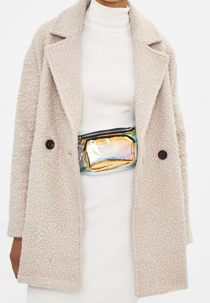 Cappotto beige con texture e colletto, chiusura con bottoni, indossato sopra un dolcevita bianco e una gonna a coste. Presenta una borsa a tracolla olografica lucida.