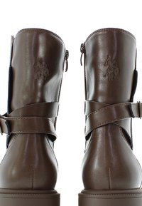 Las botas de tobillo de cuero marrón presentan un diseño de tiras cruzadas, cremalleras laterales y un acento de logo. Tienen una textura suave, con punta redondeada y un tacón grueso.