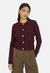 Cardigan côtelé bourgogne avec manches longues, col rond, deux poches avant et boutons dorés. Associé avec un pantalon marron.