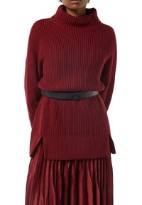Czerwony sweter z dzianiny z wysokim kołnierzem, z teksturowanym wzorem ściągacza i bocznymi rozporkami, noszony na plisowanej metalicznej spódnicy, podkreślony czarnym paskiem.