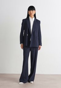 Donna in piedi all'interno che indossa un completo blu navy a doppio petto con pantaloni a gamba larga, camicia bianca, orecchini e scarpe bianche a punta su un pavimento beige.