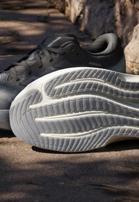 Chaussures de course noires Adidas avec des motifs visibles sur la semelle blanche, placées en extérieur sur une surface en béton.