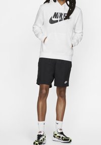 Hoodie Nike blanc avec un grand logo noir, associé à un short noir portant un petit logo. Les chaussettes sont blanches et les chaussures sont noires avec des accents néon.