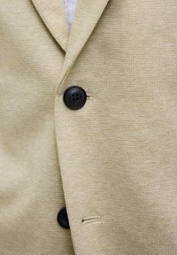 Beige blazer featuring a textured fabric with a houndstooth pattern, black buttons, and a single vent detail.