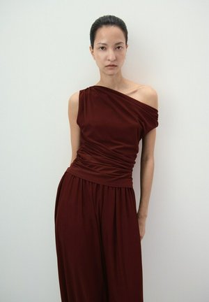 Ženska stoji ob belem zidu, oblečena v brezrokavno bordo rdečo obleko z enim ramenom, z nagubanim pasom in letečo krilo, obračena naprej z nevtralnim izrazom.