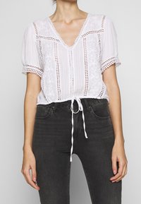 Weiße bestickte Bluse mit kurzen Ärmeln und V-Ausschnitt; verfügt über einen Kordelzugsaum und dekorative Spitzenakzente. Kombiniert mit dunklen Jeans.