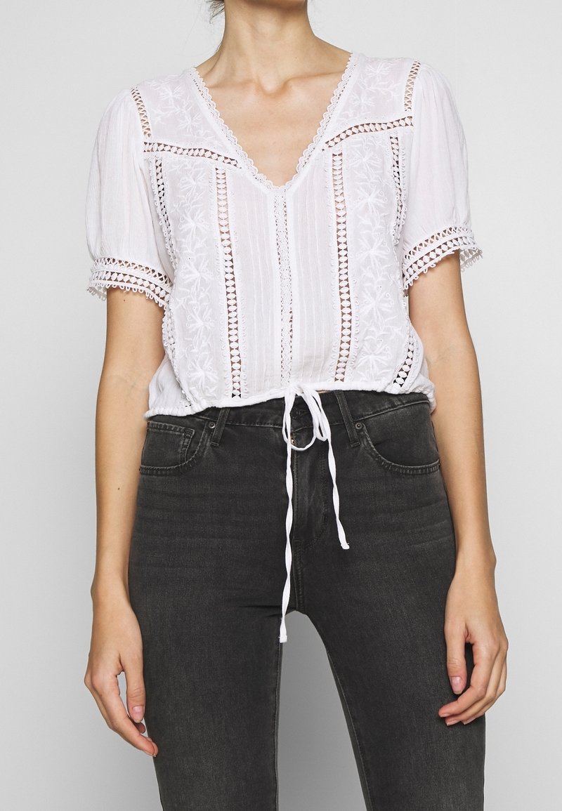 Weiße bestickte Bluse mit kurzen Ärmeln und V-Ausschnitt; verfügt über einen Kordelzugsaum und dekorative Spitzenakzente. Kombiniert mit dunklen Jeans.