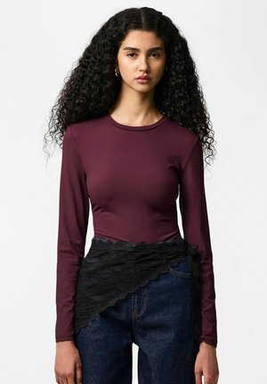 Žena s dlhými kučeravými vlasmi má na sebe bordový top s dlhým rukávom a tmavé džínsy s čiernym čipkovaným opaskom na bielom pozadí.