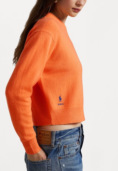 Oranž villane kampsun ribitud mansettide ja äärega, millel on sinine broderdatud logo. Kantsed kõrge vöökohaga denim lühike pükse, millel on nähtav silt.