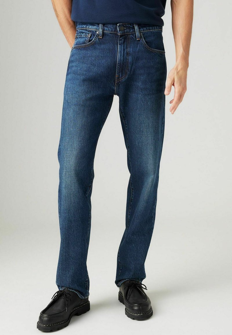 Dunkelblaue gerade geschnittene Jeans aus Denim mit einem Knopf- und Reißverschluss, Seitentaschen und dezentem Verblassen an den Oberschenkeln. Schwarz Schuhe getragen.