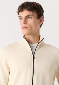 Gilet zippé beige en tricot léger avec un col montant, comportant une fermeture éclair noire et un petit logo "BOSS" sur la poitrine.