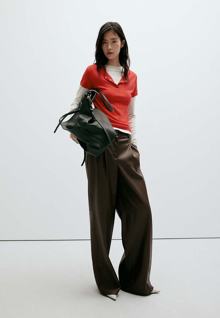 Jeune femme portant un haut rouge à manches courtes sur une chemise blanche à manches longues, pantalon large marron, tenant un grand sac en cuir noir.