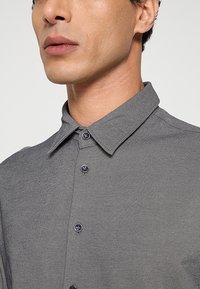 Grijs overhemd met knopen, gemaakt van een gestructureerde stof, voorzien van een puntige kraag en donkere knopen. Het ontwerp bevat subtiele diagonale strepen.