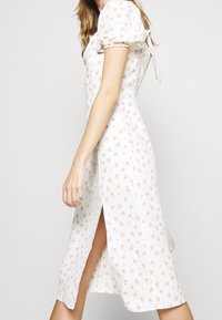 Vestido midi floral de tela blanca, con mangas abullonadas, abertura lateral y pequeñas flores rosas en todo su diseño. Se ajusta en la parte de atrás con lazo.