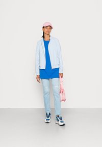 Lichtblauwe bomberjack, blauwe t-shirt, versleten lichte spijkerbroek, witte en blauwe sneakers, roze pet en een roze handtas.