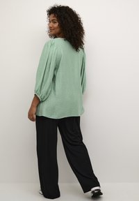 Blusa verde chiaro, con texture, maniche a sbuffo, vestibilità ampia e orlo arrotondato, abbinata a pantaloni neri a gamba larga e sneaker.