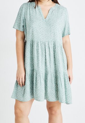 Kobieta w jasnorozowej sukience w liście z krótkim rękawem i falbankowym dołem, stojąca na jednolitym białym tle.