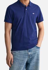 Navy blue polo shirt in cotton blend, featuring a ribbed collar and short sleeves, with a small embroidered logo on the left chest. Paired with light denim shorts.