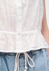 Κοντινό της λευκής υφασμάτινης αμάνικης μπλούζας με κουμπιά, συνδυασμένη με ανοιχτό μπλε τζιν παντελόνια με δεμένη κορδονένια μέση.