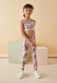 Cropped ljubičasti tie-dye set za aktivnost s prilagođenim gornjim dijelom i tajicama, izrađen od rastezljivog materijala. Bijele tenisice upotpunjuju outfit.
