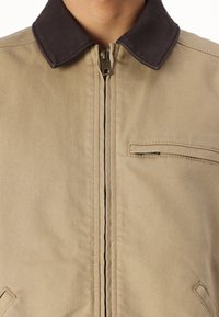 Chaqueta beige con un cuello oscuro, cierre de cremallera y un pequeño bolsillo en el pecho izquierdo. Fabricada con una tela resistente con detalles de costura visibles.