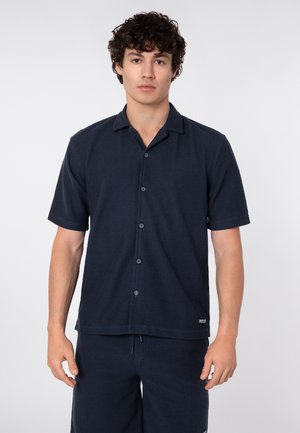 Junger Mann mit lockigem Haar, der ein dunkelblaues kurzärmeliges Hemd mit Knöpfen und passende Shorts trägt, steht vor einem einfarbigen Hintergrund.