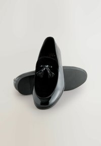 Schwarze Loafers mit Obermaterial aus glänzendem Lackleder, versehen mit einem Samtpanel und einer Schleifenakzent mit Quasten. Strukturierte Gummi-Außensohlen.