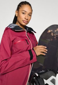 Vinrød vandtæt jakke med grå hætte, lynlåslommer og velcroer ved ærmerne, holdt af en person ved siden af et sort snowboard.