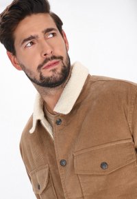 Braune Cordjacke mit einem cremefarbenen Lammfellkragen, ausgestattet mit zwei Vordertaschen und schwarzen Knopfdetails. Weicher Textur und strukturierte Passform.