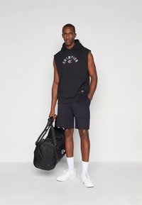 Homme portant un sweat à capuche sans manches noir et un short, chaussures et chaussettes blanches, tenant un sac de sport noir, debout devant un fond clair uni.