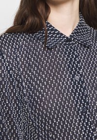 Mörkblå genomskinlig blus med ett litet prickmönster, knappstängning och spetsig krage. Mjuk textur och lätt design.