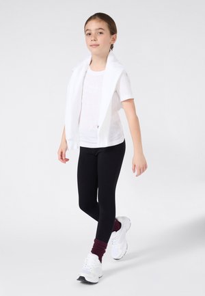 T-shirt blanc à manches courtes avec détail imprimé, leggings noirs et baskets blanches portés avec des chaussettes bordeaux mi-mollet. Fond clair.