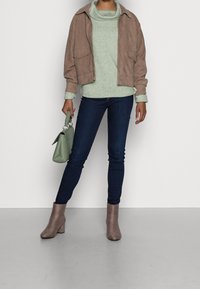 Veste courte en daim marron, pull à col bénitier vert clair, jean skinny bleu foncé, bottines à talon bloc gris, sac à main vert avec poignée tressée.