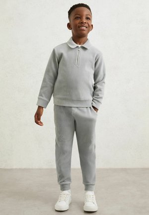 Mladý chlapec stojí, oblečený v světle šedém žebrovaném svetru na zip a odpovídajících kalhotách s bílými teniskami, usmívá se a dívá se vzhůru.