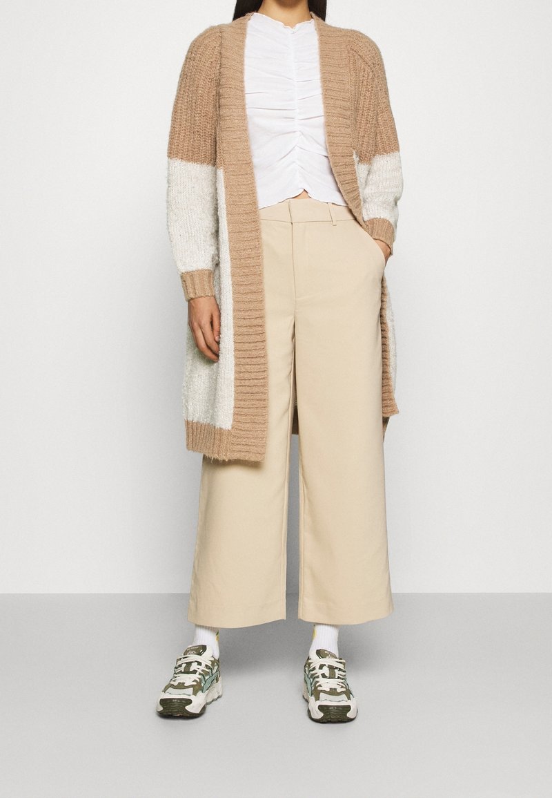 Cardigan en tricot avec des blocs de couleur beige et blanc, associé à un haut blanc froncé et un pantalon beige à jambes larges. Baskets à motif vert et blanc.