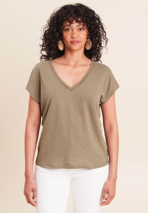 Vrouw met krullend haar, gekleed in een olijfgroene V-hals top met korte mouwen, witte broek en grote bloemige oorbellen, staand tegen een effen achtergrond.