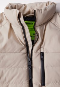 Beige gesteppte Jacke mit hohem Kragen, schwarzen Reißverschlüssen und schwarzen Akzenten; enthält ein leuchtend grünes Markenetikett im Inneren.