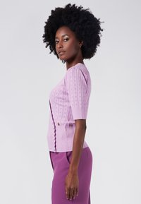 Lila stickad kofta med korta ärmar, med ett strukturerat mönster, ribbad fåll och knäppning fram. Slätt, mjukt material.