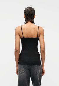 Άτομο με κοτσίδες που φοράει μαύρο τοπ με λεπτές τιράντες και φαρδιά σκούρα γκρι τζιν, εμφανιζόμενο από πίσω σε απλό φόντο.