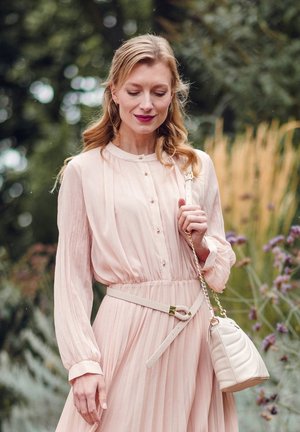 Svetle růžové sklady šaty s dlouhými rukávy, zapínáním na knoflíky a látkovým páskem. V kombinaci s krémovou kabelkou s pozlaceným řetízkem.