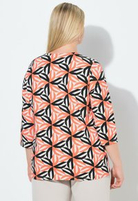 Mujer con cabello largo y rubio, vestida con una blusa de patrón geométrico en naranja, negro y blanco, con mangas tres cuartos, posando frente a una pared blanca.