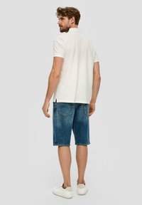 Weißes Polohemd mit kurzen Ärmeln, kombiniert mit blauen Jeansshorts. Das Model trägt weiße Sneakers. Einfaches, sauberes Design.