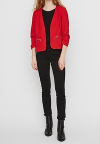 Red blazer with a textured finish, open front, and two metallic zippered pockets. Paired with a black top and slim black pants. Black ankle boots.